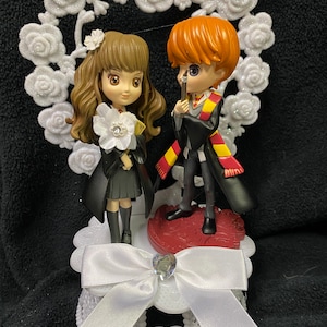 May include: A white cake topper featuring a heart-shaped rose garland. The topper depicts two figures, a girl with brown hair and a boy with red hair, both wearing black robes and holding wands. The girl is holding a white flower. The topper sits on a white base with a white bow and a rhinestone.
