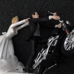 以下が含まれることがあります： 白のウェディングケーキのトッパーで、新郎新婦が描かれています。新郎は黒いオートバイに乗っており、新婦は彼に手を伸ばしています。オートバイは銀色の車輪とハンドルに赤いディテールがあります。
