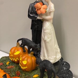 May include: A Halloween wedding cake topper with a bride and groom figurine, two carved pumpkins, and black cat figurines. The bride wears a white dress, and the groom wears a black tuxedo. The pumpkins are orange with carved faces.