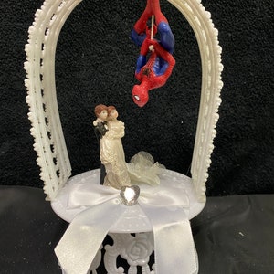 May include: White wedding cake topper with a bride and groom figurine standing under a white archway. A Spiderman figurine is hanging upside down from the archway.