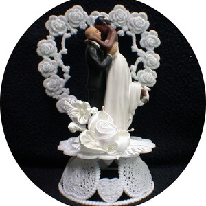 May include: White wedding cake topper featuring a bride and groom embracing. The bride is wearing a white dress and the groom is wearing a black suit. The couple is standing in front of a white heart-shaped frame decorated with glitter.
