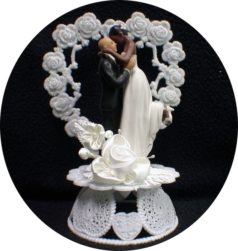 May include: White wedding cake topper featuring a bride and groom embracing. The bride is wearing a white dress and the groom is wearing a black suit. The couple is standing in front of a white heart-shaped frame decorated with glitter.