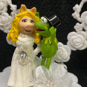 Puede incluir: Un adorno para pastel de boda blanco con una rana verde que lleva un sombrero de copa negro y un cerdo que lleva un vestido de novia blanco.
