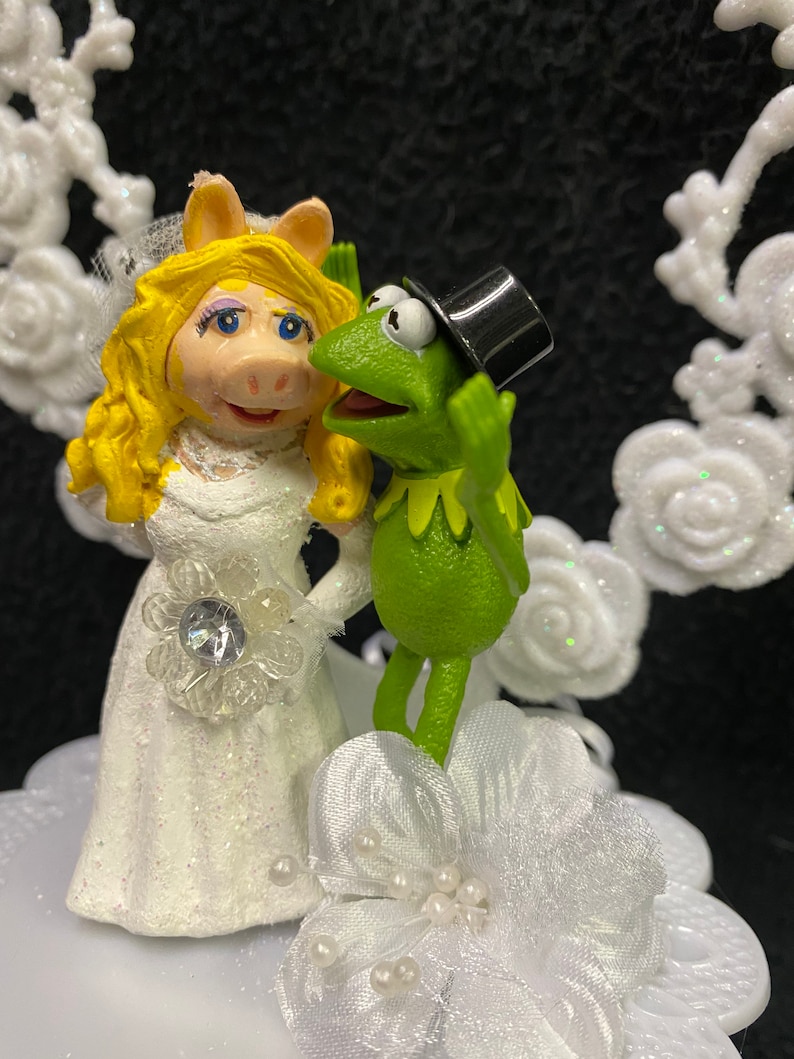 Puede incluir: Un adorno para pastel de boda blanco con una rana verde que lleva un sombrero de copa negro y un cerdo que lleva un vestido de novia blanco.