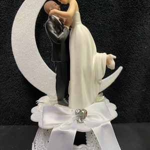 Pu&ograve; includere: Un topper per torta bianco con una sposa e uno sposo che si abbracciano su una luna crescente bianca. La sposa indossa un abito bianco e lo sposo indossa uno smoking nero. La base del topper &egrave; bianca con un fiocco bianco e un cuore di strass.