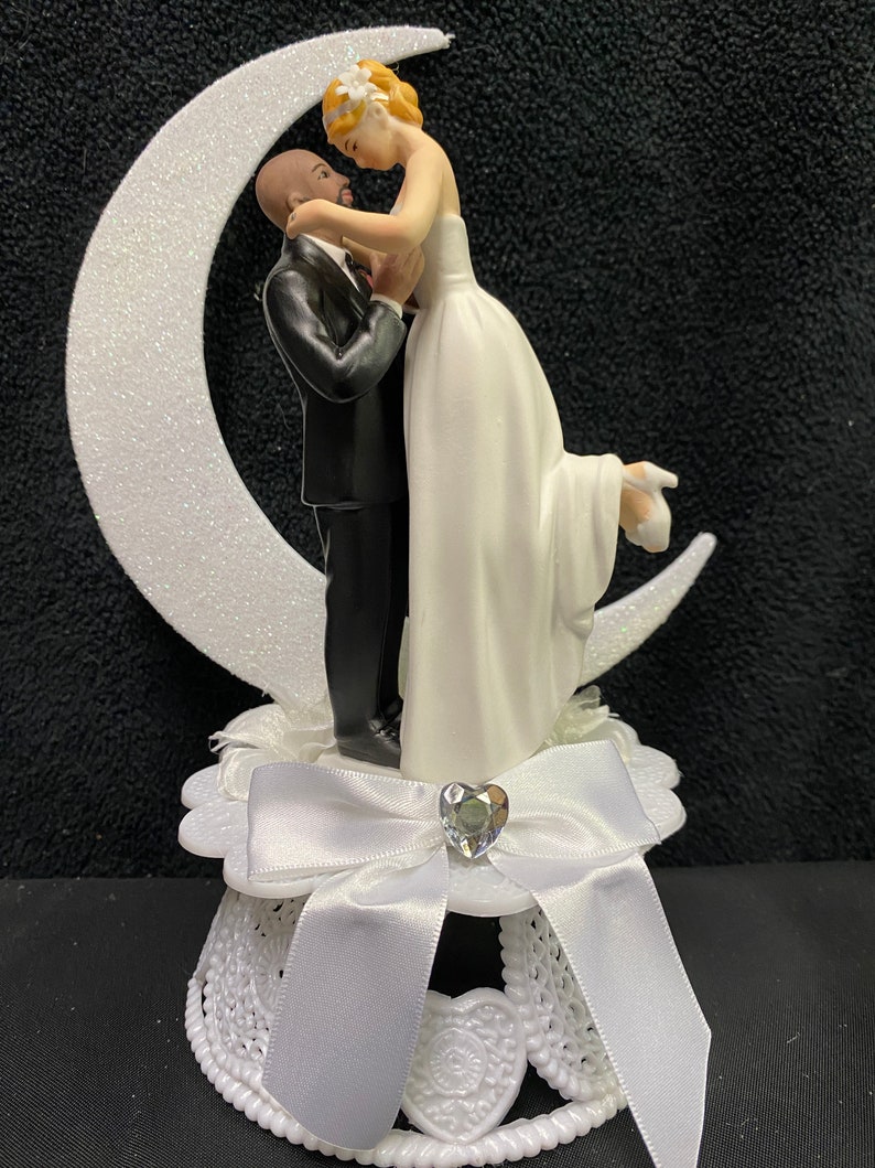 Pu&ograve; includere: Un topper per torta bianco con una sposa e uno sposo che si abbracciano su una luna crescente bianca. La sposa indossa un abito bianco e lo sposo indossa uno smoking nero. La base del topper &egrave; bianca con un fiocco bianco e un cuore di strass.
