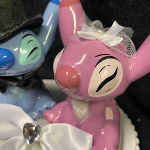 May include: A wedding cake topper featuring two cartoon characters. One is blue and wears a black top hat and bow tie. The other is pink and wears a white veil and bow. A white bow with a heart-shaped gem is in the foreground.