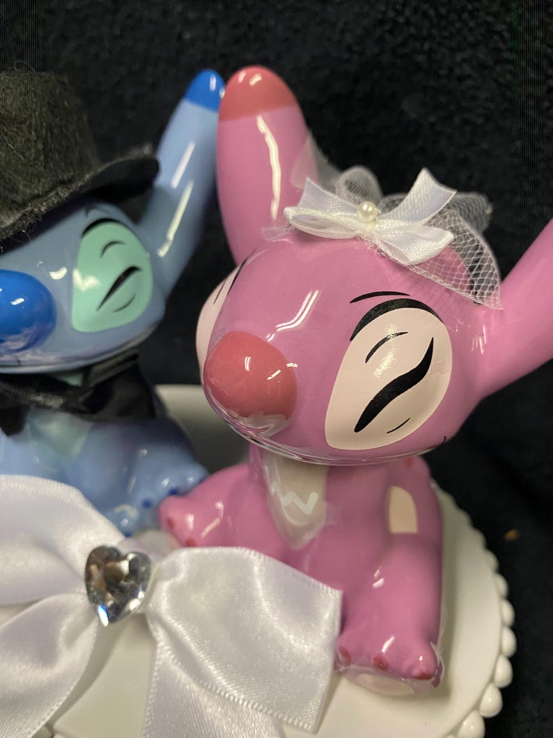 May include: A wedding cake topper featuring two cartoon characters. One is blue and wears a black top hat and bow tie. The other is pink and wears a white veil and bow. A white bow with a heart-shaped gem is in the foreground.