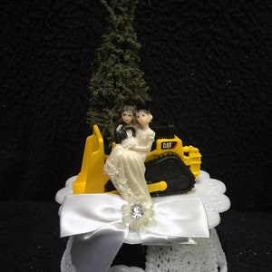 Op de afbeelding: Een geel speelgoed-bouwvoertuig met een bruidspaarfiguur erop. Het voertuig is gemarkeerd met "CAT" en staat op een witte taarttopper met een witte strik en een strasssteen.