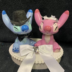 May include: A wedding cake topper with two cartoon figures, one blue and one pink, with black bow ties and a white bow. The blue figure wears a black top hat. The topper sits on a white base with a decorative edge.
