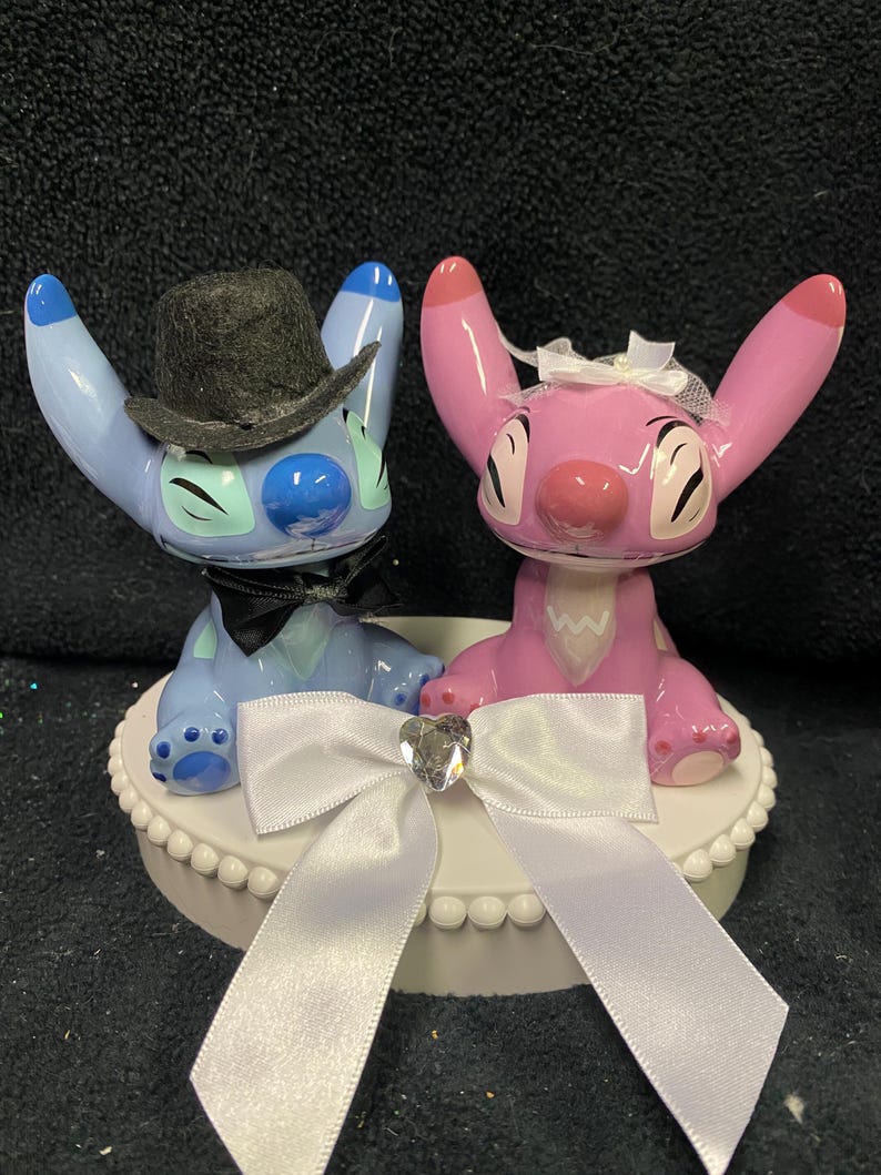 May include: A wedding cake topper with two cartoon figures, one blue and one pink, with black bow ties and a white bow. The blue figure wears a black top hat. The topper sits on a white base with a decorative edge.