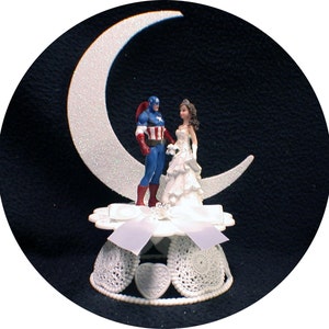 May include: A white cake topper featuring a bride and groom figurine standing on a crescent moon. The groom is dressed as Captain America, wearing a blue and red costume with a star on his chest. The bride is wearing a white gown with a tiara. The cake topper is on a white base with a white ribbon and lace trim.