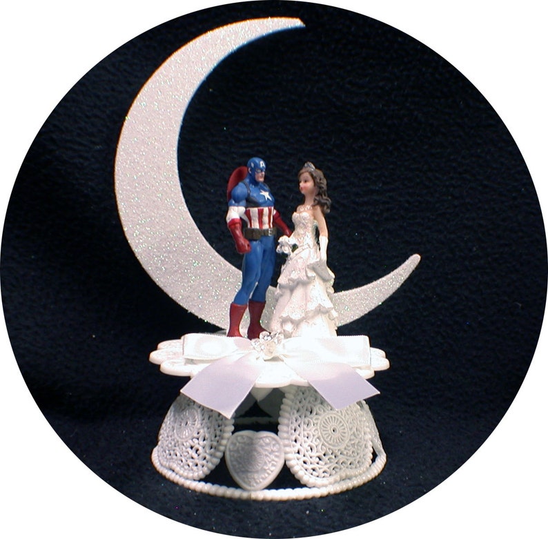 May include: A white cake topper featuring a bride and groom figurine standing on a crescent moon. The groom is dressed as Captain America, wearing a blue and red costume with a star on his chest. The bride is wearing a white gown with a tiara. The cake topper is on a white base with a white ribbon and lace trim.