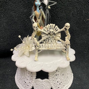 Puede incluir: Un adorno para pastel blanco con Jack Skellington y Sally de Pesadilla Antes de Navidad. Están de pie sobre un trono esquelético con una base blanca.