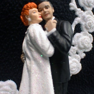 May include: A wedding cake topper featuring a bride and groom in a formal pose. The bride is wearing a white dress with glitter and the groom is wearing a black tuxedo. The couple is surrounded by white, sparkly decorations.