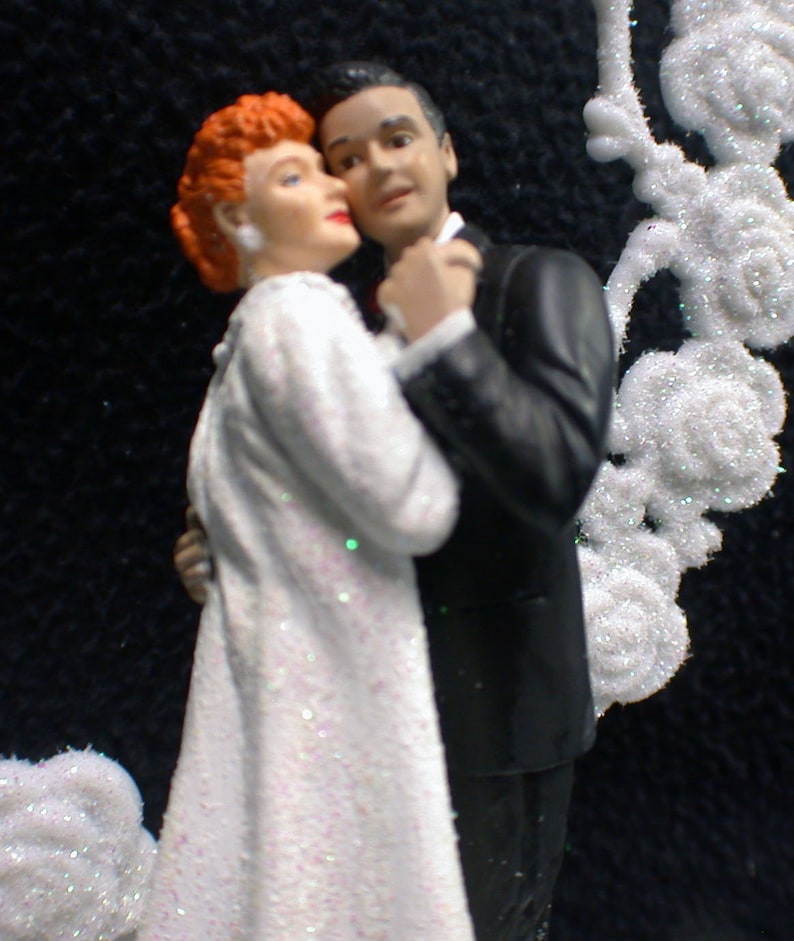 May include: A wedding cake topper featuring a bride and groom in a formal pose. The bride is wearing a white dress with glitter and the groom is wearing a black tuxedo. The couple is surrounded by white, sparkly decorations.