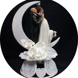 May include: A white and black wedding cake topper featuring a bride and groom kissing on a crescent moon. The bride is wearing a white dress and the groom is wearing a black tuxedo. The base of the topper is decorated with white flowers and leaves.