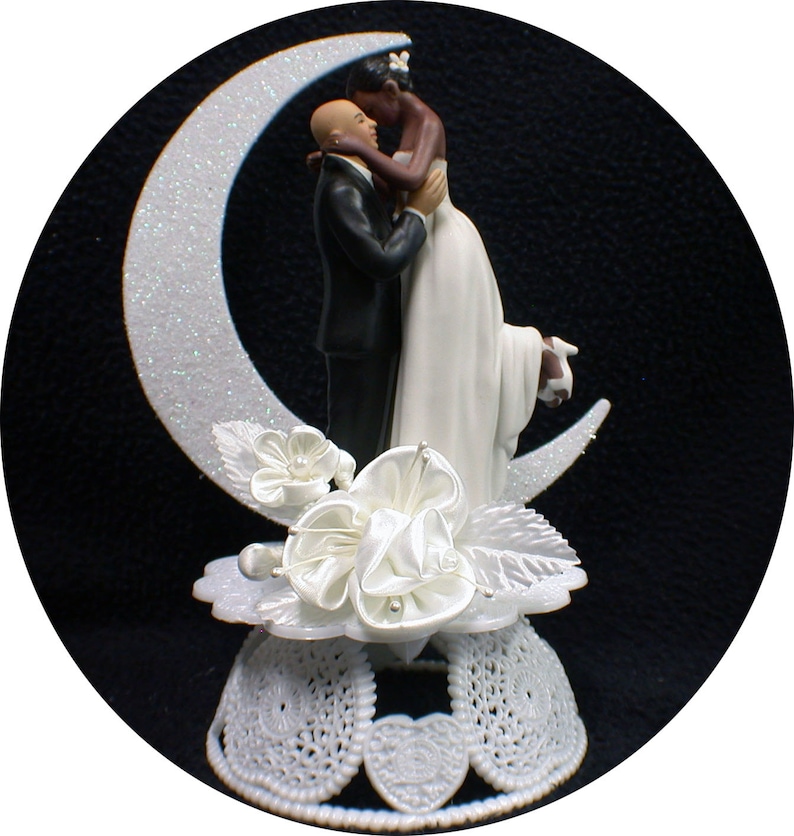May include: A white and black wedding cake topper featuring a bride and groom kissing on a crescent moon. The bride is wearing a white dress and the groom is wearing a black tuxedo. The base of the topper is decorated with white flowers and leaves.