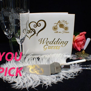 May include: Wedding cake cutting set with a silver knife and a white handle. The knife has a gold design of two intertwined hearts. The set is on a white feather boa with a white guest book and two champagne flutes in the background. The guest book has a gold design of a motorcycle with the text "Ride with me Forever" and "Wedding Guests".