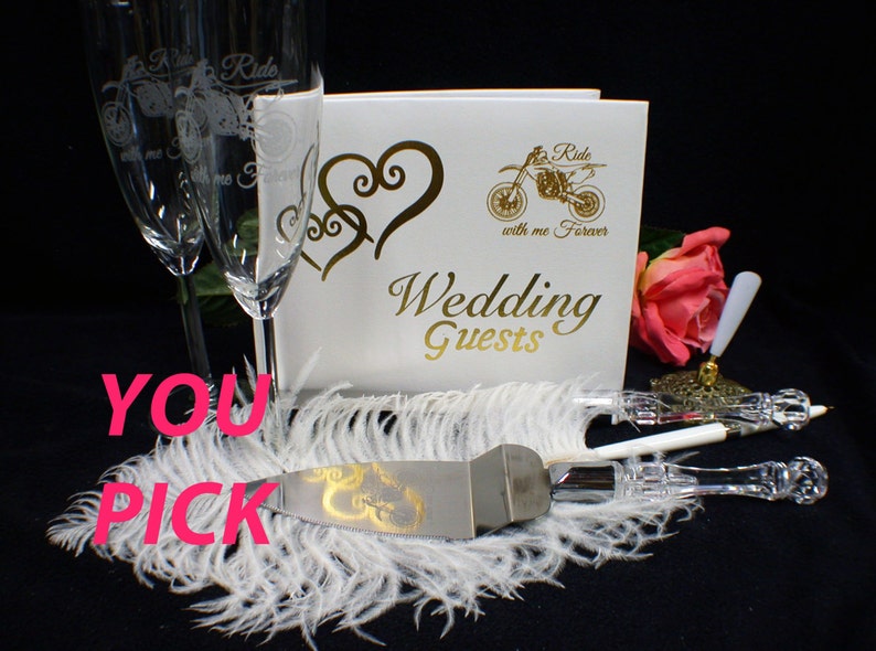 May include: Wedding cake cutting set with a silver knife and a white handle. The knife has a gold design of two intertwined hearts. The set is on a white feather boa with a white guest book and two champagne flutes in the background. The guest book has a gold design of a motorcycle with the text "Ride with me Forever" and "Wedding Guests".