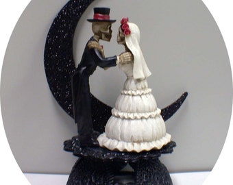 May include: A black and white skeleton wedding cake topper with the bride and groom standing on a crescent moon. The bride is wearing a white dress with a veil and a red flower crown. The groom is wearing a black suit and a top hat.