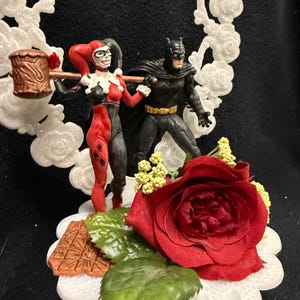 May include: A cake topper featuring miniature figures of Harley Quinn holding a brown mallet and Batman standing beside her. The figures are set against a white, glittery, rose-patterned circular frame, accented with a large red rose and green leaves.