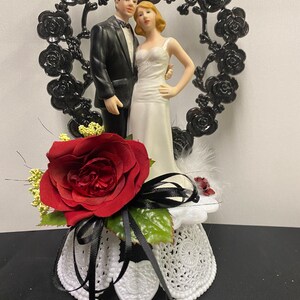 May include: A black and white wedding cake topper featuring a bride and groom standing in front of a black heart-shaped frame with black roses. The base of the topper is white with a black ribbon and a red rose.