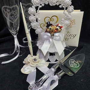 Może przedstawiać: Biały topper na tort ślubny z kreskówkową parą w ramce w kształcie serca. Para stoi na białej podstawie tortu z białą kokardką. Topper na tort jest otoczony białymi różami. Do zestawu należą dwa kieliszki do szampana, stojak na długopis i nóż do tortu. Nóż do tortu ma kształt serca z kwiatowym wzorem.