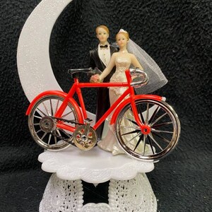 Puede incluir: Un adorno de pastel de boda de bicicleta roja con una figura de novia y novio. La novia lleva un vestido blanco y velo, y el novio lleva un esmoquin negro. La bicicleta está sobre una base blanca con una luna creciente detrás.