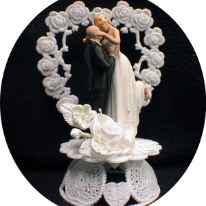 May include: White cake topper featuring a bride and groom figurine. The bride is wearing a white dress and the groom is wearing a black tuxedo. The figurine is standing on a white base with a heart-shaped design.