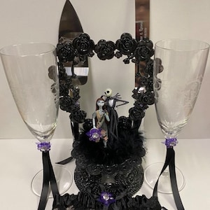Może przedstawiać: Czarny topper na tort weselny z Jackiem Skellingtonem i Sally z filmu Koszmar przed świętami. Topper stoi na czarnym stojaku na tort z czarnymi różami i czarną i białą podwiązką. W zestawie dwie fluty do szampana z fioletowymi wstążkami i zawieszkami.