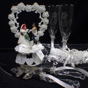 Może przedstawiać: Biały topper na tort przedstawiający parę młodą podczas ceremonii ślubnej. Panna młoda ma na sobie białą suknię i czerwoną ozdobę do włosów. Pan młody ma na sobie niebieski garnitur. Topper na tort jest otoczony białą, sercową ramą z białymi różami. W zestawie znajdują się dwa kieliszki do szampana z wzorem pary młodej. W zestawie znajduje się również biała podwiązka z koronką i białą wstążką. W zestawie znajduje się srebrny nóż do tortu z kryształową rączką.