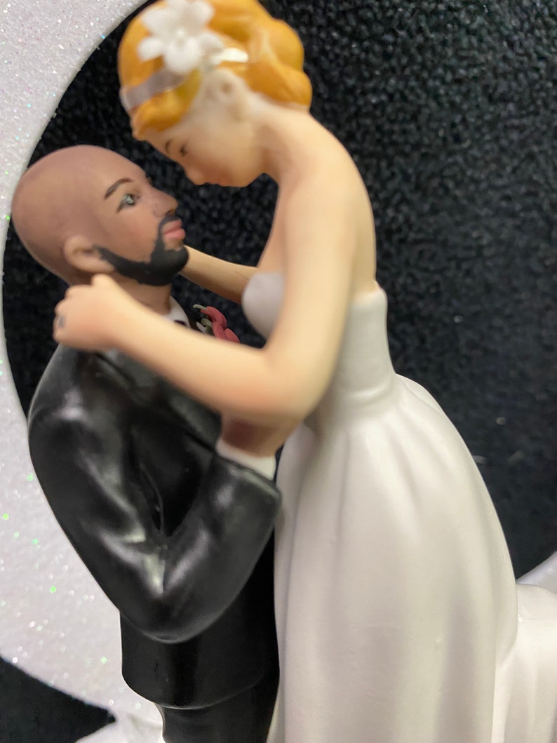 Pu&ograve; includere: Un topper per torta nuziale con una sposa e uno sposo. La sposa indossa un abito bianco e lo sposo indossa un abito nero. La sposa tiene il viso dello sposo e si guardano.