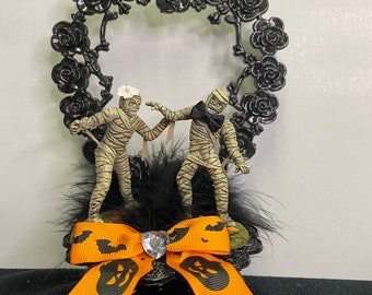 May include: A black metal heart-shaped cake topper with a black and white mummy bride and groom figurine. The topper is decorated with black feathers and an orange and black bow with a rhinestone in the center.