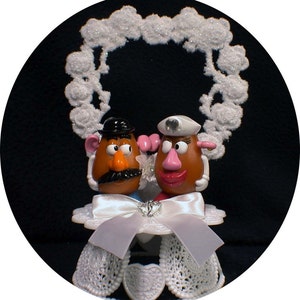 Puede incluir: Un adorno para pastel blanco con Mr. y Mrs. Potato Head, vestidos con atuendos de boda, de pie bajo un marco blanco en forma de corazón. El adorno está sobre una base blanca con un borde de encaje blanco y un lazo de satén blanco.