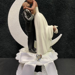 Pu&ograve; includere: Un topper per torta bianco con una sposa e uno sposo che si abbracciano su una luna crescente. La sposa indossa un abito bianco e lo sposo indossa un abito nero. La base del topper &egrave; bianca con un fiocco bianco e un cuore di strass.