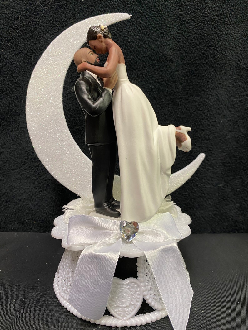 Pu&ograve; includere: Un topper per torta bianco con una sposa e uno sposo che si abbracciano su una luna crescente. La sposa indossa un abito bianco e lo sposo indossa un abito nero. La base del topper &egrave; bianca con un fiocco bianco e un cuore di strass.