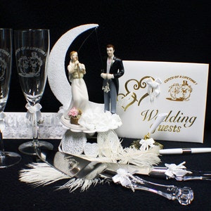 Könnte beinhalten: Ein weißer Hochzeitstortenaufsatz mit einer Braut und einem Bräutigam, die von einem Halbmond aus angeln. Der Tortenaufsatz wird von zwei Champagnerflöten, einem Tortenmesser und einem weißen Gästebuch mit dem Text "Catch of a Lifetime! Wedding Guests" begleitet.
