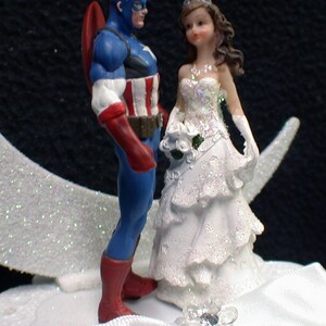 May include: A wedding cake topper featuring a bride and groom figurine. The groom is dressed as Captain America, wearing a blue and red costume with a star on his chest. The bride is wearing a white gown with a tiara and a bouquet of flowers.