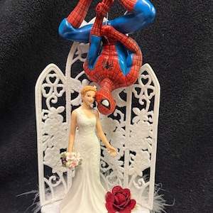 May include: A wedding cake topper featuring a bride in a white gown holding a bouquet, standing before a white decorative lattice screen. Above the bride, a red and blue Spider-Man figure hangs upside down. A single red rose sits at the base.