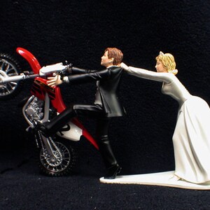 May include: A red and white motocross bike with a groom figurine holding onto the handlebars and a bride figurine pulling the groom away from the bike.