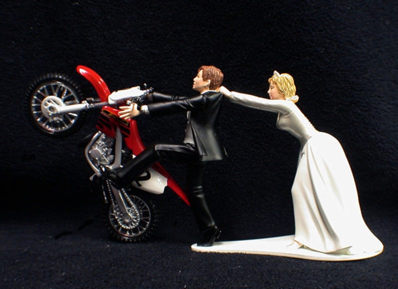 May include: A red and white motocross bike with a groom figurine holding onto the handlebars and a bride figurine pulling the groom away from the bike.