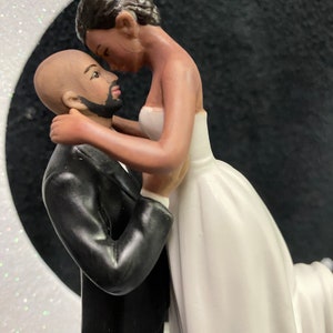 Pu&ograve; includere: Un topper per torta nuziale in bianco e nero con una sposa e uno sposo che si abbracciano. La sposa indossa un abito bianco e lo sposo indossa uno smoking nero. La coppia &egrave; in piedi su una mezzaluna.