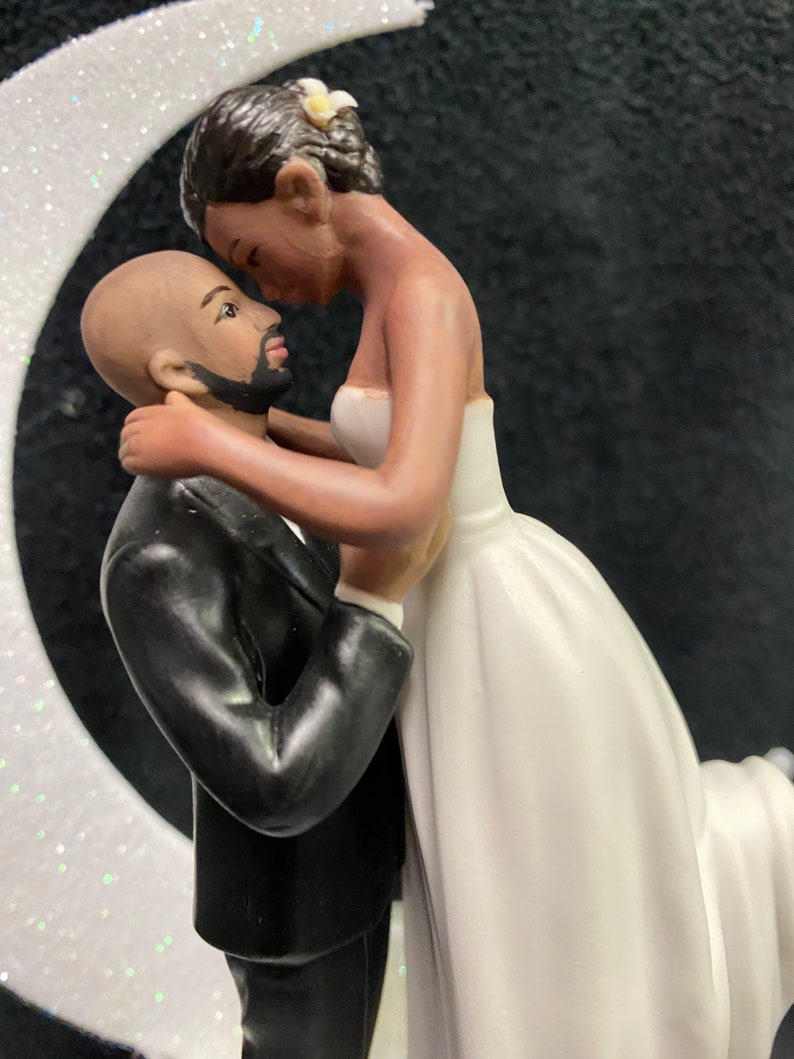 Pu&ograve; includere: Un topper per torta nuziale in bianco e nero con una sposa e uno sposo che si abbracciano. La sposa indossa un abito bianco e lo sposo indossa uno smoking nero. La coppia &egrave; in piedi su una mezzaluna.