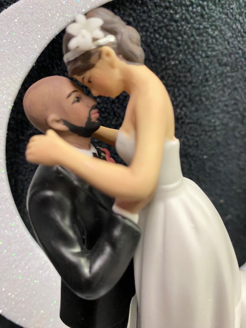 Pu&ograve; includere: Un topper per torta nuziale con una sposa e uno sposo che si abbracciano. La sposa indossa un abito bianco e lo sposo indossa un abito nero. La coppia &egrave; in una posa di abbraccio romantico.