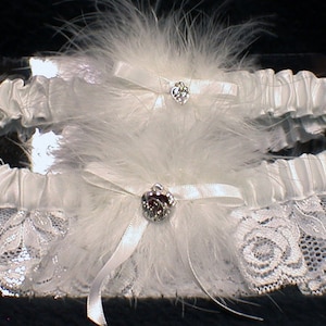 May include: A pair of white bridal garters with lace and satin. Each garter has a white feather boa and a rhinestone heart-shaped embellishment.