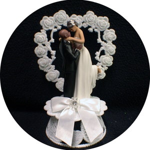 Puede incluir: Un adorno de pastel blanco con una novia y un novio abraz&aacute;ndose. La novia lleva un vestido blanco y el novio lleva un esmoquin negro. La pareja est&aacute; rodeada de un dise&ntilde;o floral blanco en forma de coraz&oacute;n.