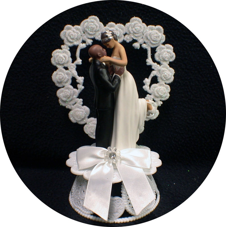 Puede incluir: Un adorno de pastel blanco con una novia y un novio abraz&aacute;ndose. La novia lleva un vestido blanco y el novio lleva un esmoquin negro. La pareja est&aacute; rodeada de un dise&ntilde;o floral blanco en forma de coraz&oacute;n.