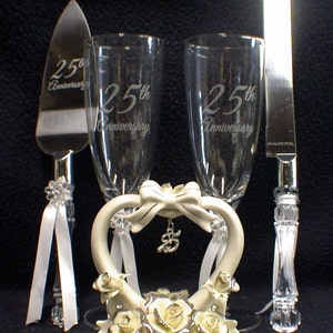 Könnte beinhalten: Ein 25. Hochzeitstag-Kuchentopper-Set mit zwei Champagnerflöten, einem Kuchmesser und einer herzförmigen Keramikbasis mit Rosen und einer Schleife. Die Flöten und das Messer sind mit "25. Hochzeitstag" graviert.