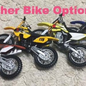 May include: Four toy dirt bikes in different colors, including orange, yellow, and blue. Each bike has a number "119" printed on it. The bikes are arranged in a row on a brown surface. The text "Other Bike Options" is written in pink above the bikes.